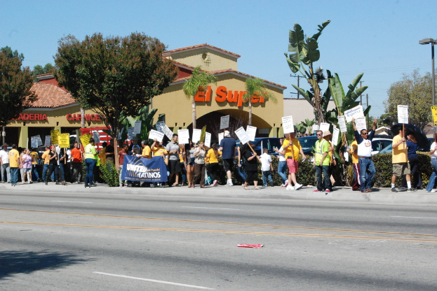 Eine Gruppe von Menschen mit Schildern und Bäumen, kleinen Büschen, einer Fahne, einem Gebäude mit Namensschildern und Autos im Hintergrund, möglicherweise während eines aktuellen Umfrageereignisses.