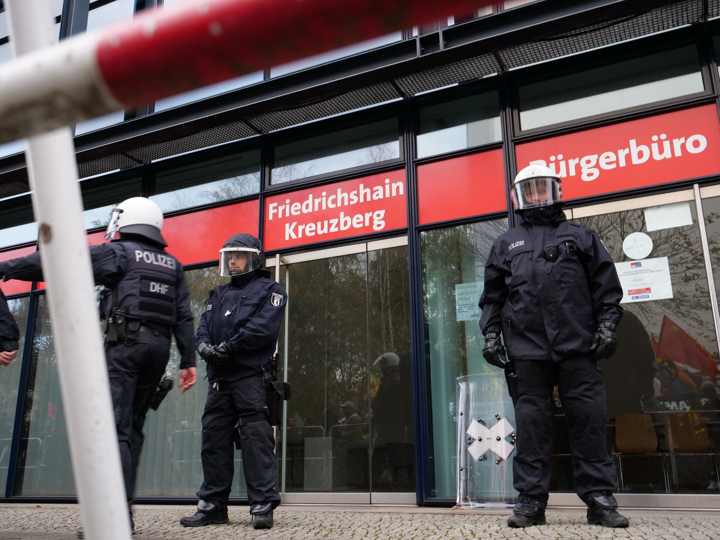 Polizisten in Helmen und Handschuhen stehen vor einem Gebäude mit Glaswänden und -türen, neben einem Pfahl, mit Schildern, die "Friedrichshain Kreuzberg" lesen.