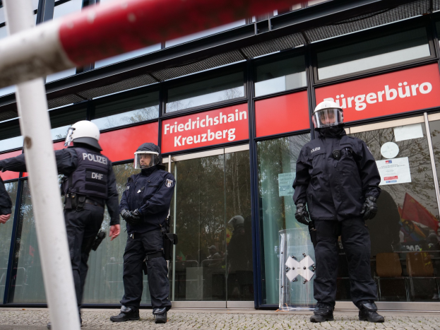 Polizisten in Helmen und Handschuhen stehen vor einem Gebäude mit Glaswänden und -türen, neben einem Pfahl, mit Schildern, die "Friedrichshain Kreuzberg" lesen.