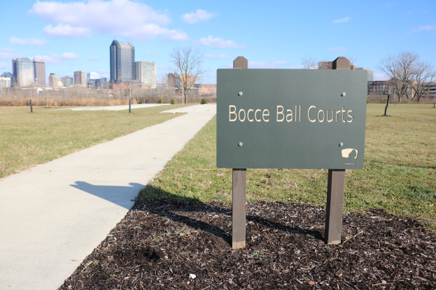 Ein Schild am Boden trägt die Aufschrift "Bocce Ball Courts in Indianapolis, Indiana" in der Nähe einer grünen Fläche mit Bäumen, einem Pfad und Gebäuden unter einem bewölkten Himmel.