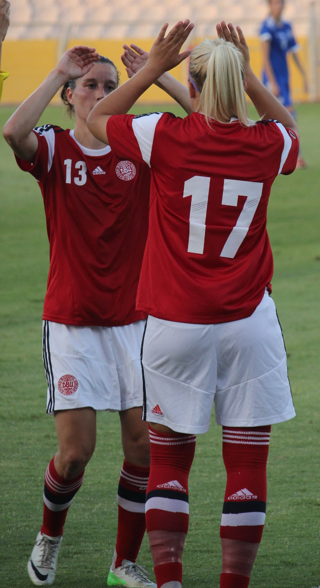 Zwei Frauen in roten T-Shirts und weißen Shorts stehen auf einem Fußballfeld und klatschen begeistert, im Hintergrund ist eine Person in einem blauen Kleid, eine gelbe Tafel und einige Stühle zu sehen.