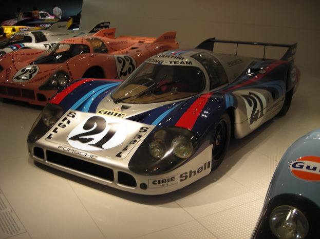 Ein Porsche 917K Rennwagen aus Le Mans in einem Museum mit anderen Sportwagen, Menschen, einer Wand im Hintergrund und einem Textschild auf der linken Seite.