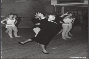 Eine Gruppe von Frauen tanzt in einem Schwarz-Weiß-Foto, wobei die zentrale Frau Tanzschuhe trägt, in einem Studio mit einer Wand im Hintergrund.