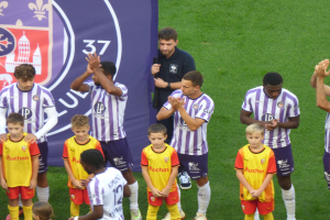 Eine Gruppe junger Jungs steht auf einem Fußballfeld, klatscht in die Hände, mit einem Banner und Logos im Hintergrund und saftigem grünem Gras unter den Füßen.