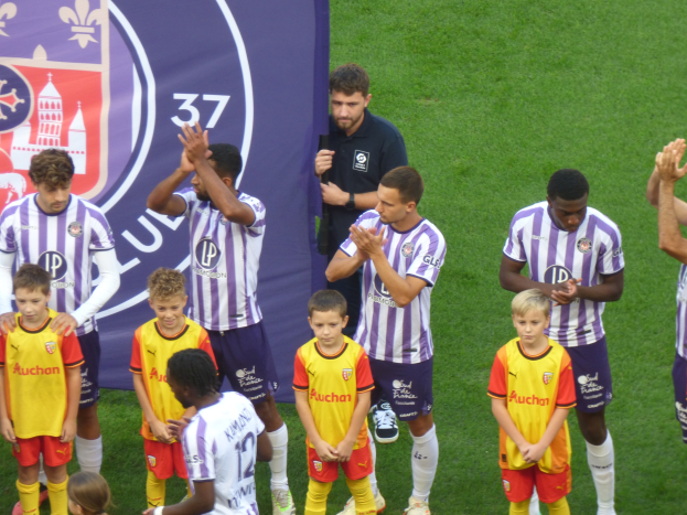 Eine Gruppe junger Jungs steht auf einem Fußballfeld, klatscht in die Hände, mit einem Banner und Logos im Hintergrund und saftigem grünem Gras unter den Füßen.