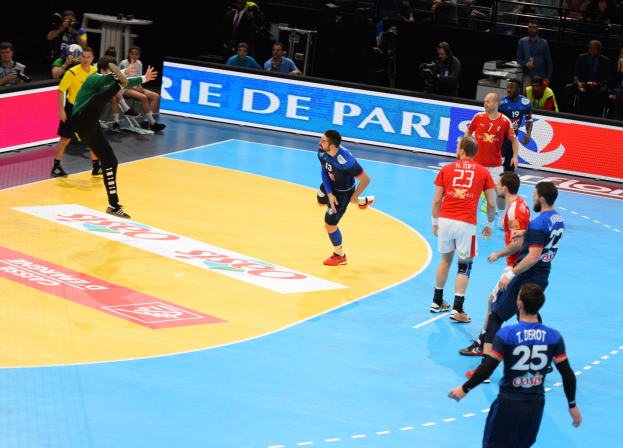 Eine Gruppe von Männern, die Handball auf einem Court spielen, mit Zuschauern und Kameras im Hintergrund und Tafeln mit Text, die auf die Futsal-Weltmeisterschaft 2019 hinweisen.