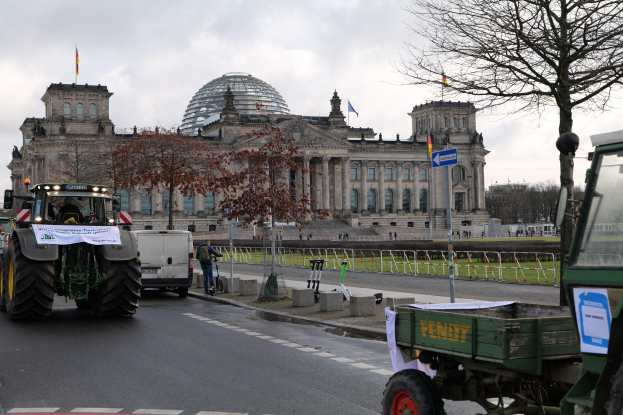 Eine Gruppe von Traktoren fährt vor dem Reichstaggebäude in Berlin, Deutschland, vorbei, das mit Fenstern, Säulen und Flaggen geschmückt ist und von Bäumen, Geländern, Pfosten und Brettern umgeben ist, mit ein paar Menschen im Hintergrund und einem bewölkten Himmel.