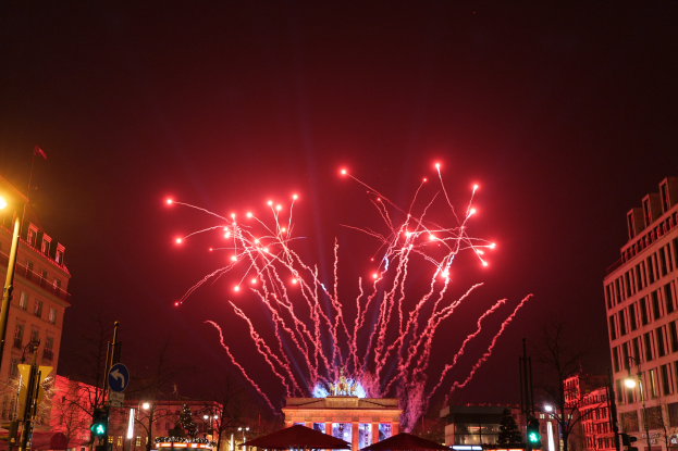 Eine Stadtstraße in Berlin an Silvester, voller Gebäude, Bäume, Laternen, Verkehrssignale, Schilder, Zelte und Menschen, mit einem Himmel, der von Feuerwerk erhellt wird.