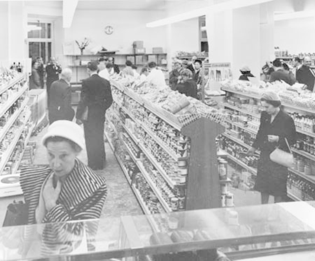 Schwarzes und weißes Foto von Menschen beim Einkaufen in einem Supermarkt mit Regalen voller Lebensmittel, einige halten Taschen, eine Uhr an der Wand und Deckenlampen.