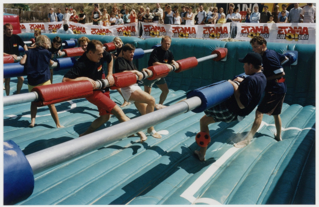 Eine Gruppe von Menschen auf einer aufblasbaren Wasserrutsche, einige halten Gegenstände, mit einem Banner, Bäumen und einem klaren blauen Himmel im Hintergrund.