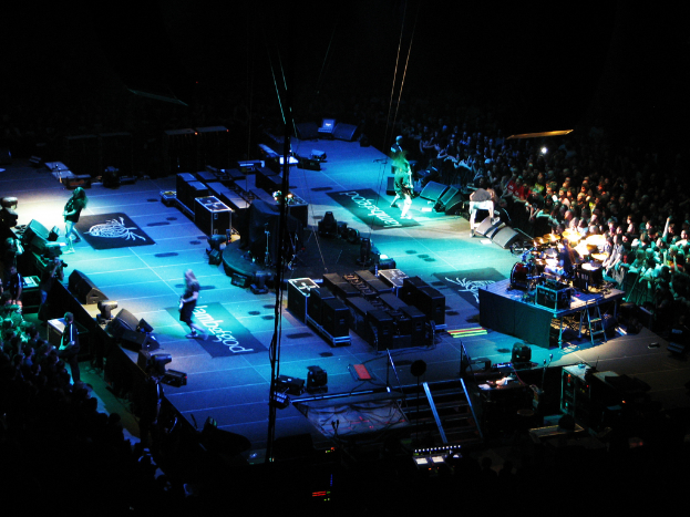 Eine Gruppe von Menschen, die auf einer Bühne mit Lautsprechern, Scheinwerfern und Mikrofonen performs, umgeben von einer großen Menge in einer dunklen Umgebung.