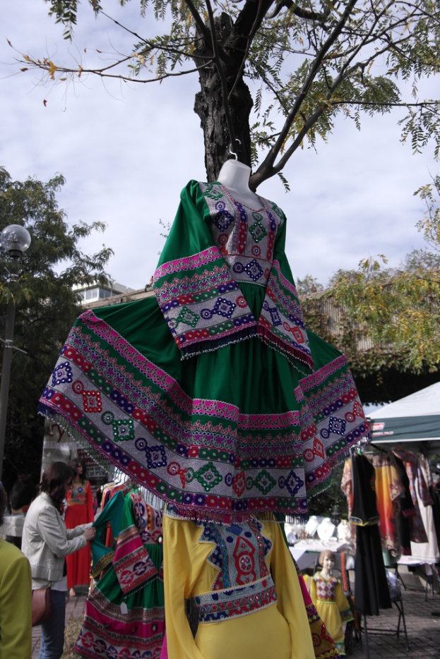 Eine Schaufensterpuppe steht in der Mitte eines belebten Einkaufsgebiets mit verschiedenen Geschäften im Hintergrund, einer Person, die Kleider an einer Stange arrangiert, einem Gebäude und Bäumen daneben.