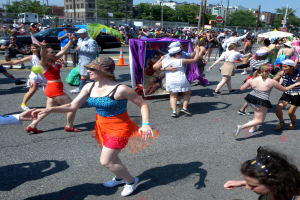 Eine Gruppe von Menschen, die auf einer Straße in einer Parade marschieren, einige mit Mützen und Schirmen, andere tanzend, mit einem Banner, Verkehrskegeln, Fahrzeugen, Laternenmasten, Schildern, Bäumen, Gebäuden und einem klaren blauen Himmel im Hintergrund.