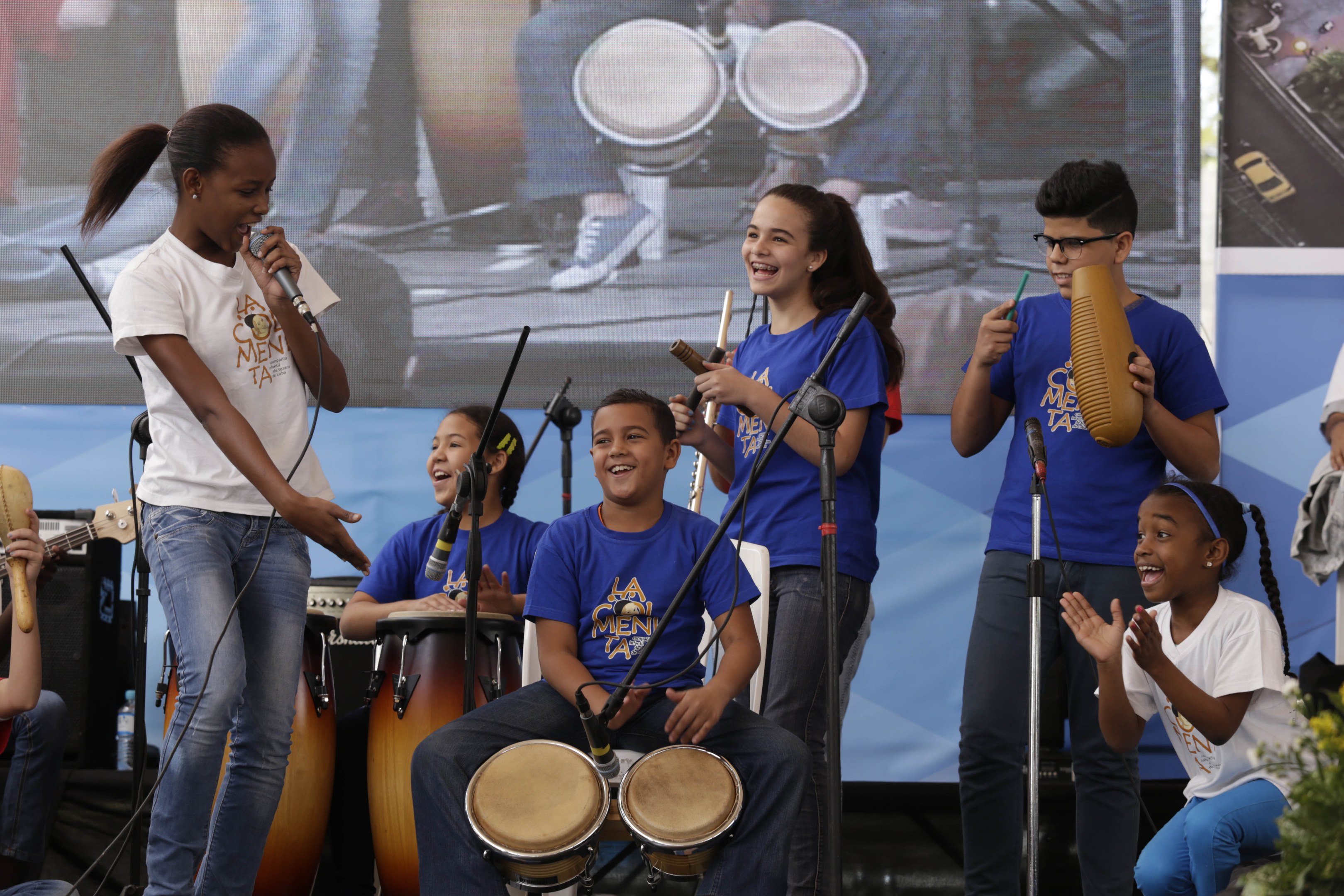 Eine Gruppe von Kindern, die auf einer Bühne Musikinstrumente spielen, mit einer Frau, die ein Mikrofon hält, in der Mitte, umgeben von Mikrofonstativen, einem Banner im Hintergrund und Pflanzen auf der rechten Seite.