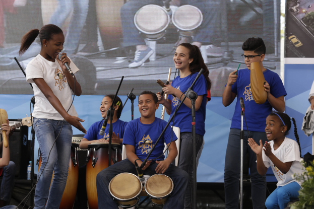 Eine Gruppe von Kindern, die auf einer Bühne Musikinstrumente spielen, mit einer Frau, die ein Mikrofon hält, in der Mitte, umgeben von Mikrofonstativen, einem Banner im Hintergrund und Pflanzen auf der rechten Seite.
