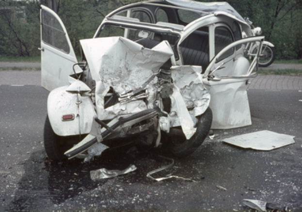 Ein Auto mit schwerer Frontschädigung steht am Straßenrand, mit Bäumen, einem Gebäude und einem anderen Fahrzeug im Hintergrund.