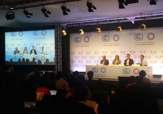 Menschen sitzen auf Stühlen auf einer Bühne mit Namensschildern und Mikrofonen auf einem Tisch, Zuschauer sitzen ihnen gegenüber, ein Projektionsschirm und Beleuchtung an Eisenstangen im Hintergrund.