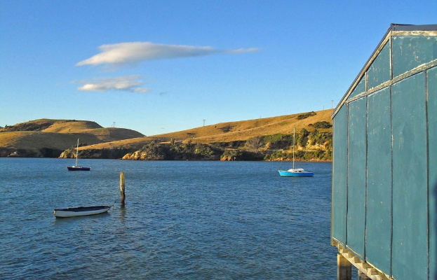 Boote und Masten im Wasser, ein Holzhaus rechts, Bäume und Berge mit Windrädern im Hintergrund und Wolken am Himmel.
