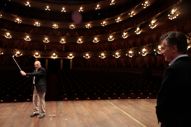 Ein älterer Mann im Anzug hält einen Golfschläger auf der Bühne, neben ihm steht ein weiterer älterer Mann, unter Deckenlampen mit vielen Sitzplätzen darunter.