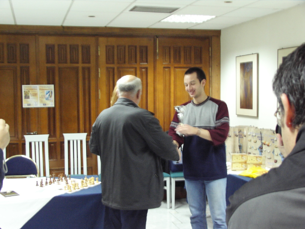 Ein Besprechungsraum mit Menschen, die stehen und sprechen, ein blauer Tisch mit einem Schachbrett und einem Stuhl davor, Schränke im Hintergrund und Fotorahmen an der rechten Wand.