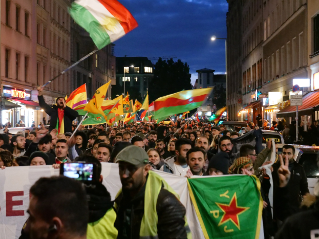 Eine große Menschenmenge marschiert mit Protestschildern durch eine Stadtstraße, mit einer Person im Vordergrund, die ein Handy verwendet, und Gebäuden, Fahrzeugen und Bäumen im Hintergrund.