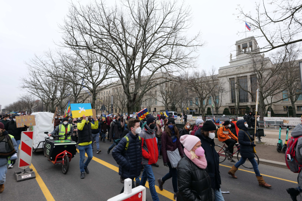 Eine große Gruppe von Menschen bei einer Demonstration auf einer Straße, einige halten Schilder und andere fahren Fahrráder, mit Bäumen und einem Gebäude im Hintergrund bei klarem blauem Himmel.