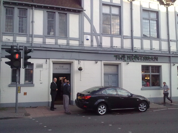 Ein Gebäude mit Text und Rohren an der Wand, ein Auto auf der Straße im Vordergrund, drei stehende Personen, ein Mann auf dem Gehweg, eine Gruppe von Personen hinter einem Fenster und ein Verkehrspfahl auf der linken Seite des Gehwegs.
