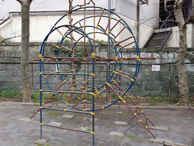 Ein Spielplatz mit einer großen Metallstruktur in der Mitte, umgeben von Bäumen und einer Wand mit Maschendrahtzaun, mit Treppen mit Geländern, die zu einem Gebäude mit Fenstern und Rohren führen, und einem Schild an einem Pfahl, alles auf einem grasbewachsenen Boden.