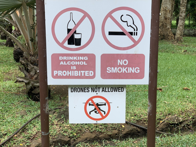 Schilder am Straßenrand mit zwei Verboten: "Alkoholtrinken ist verboten" und "Kein Rauchen", mit Bäumen und Gras im Hintergrund.