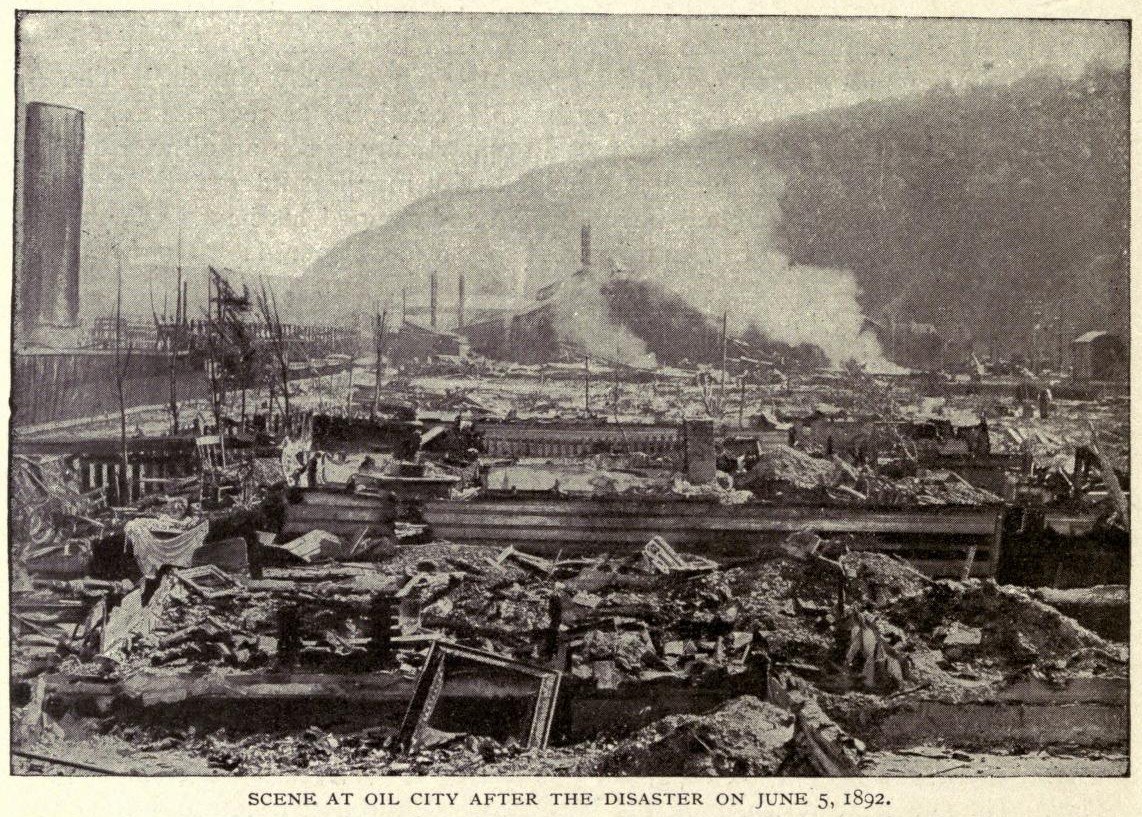 Schwarz-weiß-Foto einer Stadt nach einer Katastrophe am 5. Juni 1892, das Gebäude, Pfähle, Rauch und einen Hügel im Hintergrund zeigt, mit Text unten.