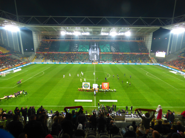 Ein großes, hell erleuchtetes Stadion voller Zuschauer, die einem Fußballspiel zusehen, mit Fans, die auf den Tribünen sitzen und um das Feld herumstehen.
