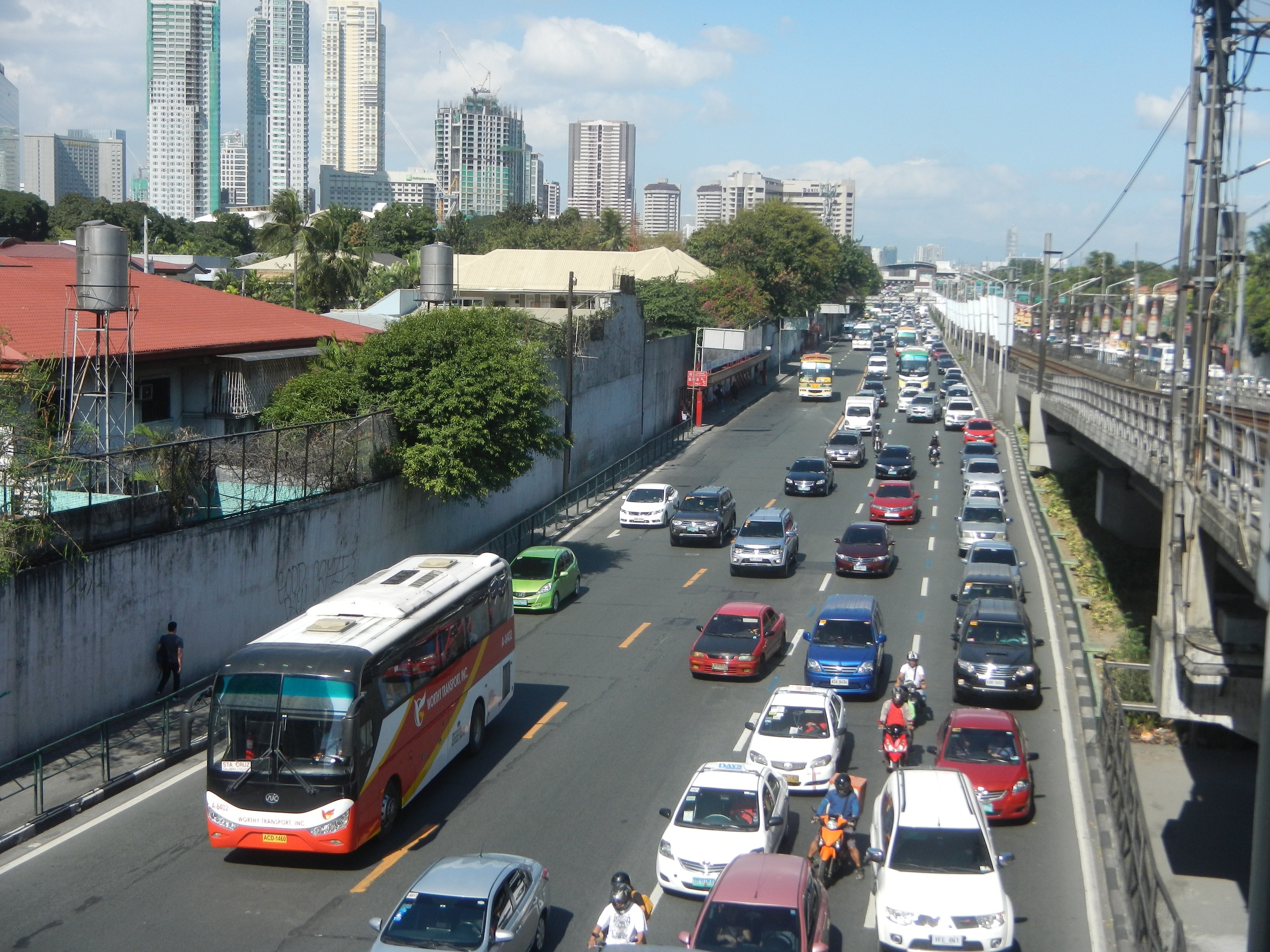 Stadtstraße mit viel Verkehr, darunter Autos und Busse, sowie Motorradfahrer, Strommasten und -leitungen auf der rechten Seite, Gebäude, Bäume und ein bewölkter Himmel im Hintergrund.