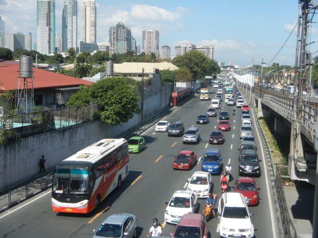 Stadtstraße mit viel Verkehr, darunter Autos und Busse, sowie Motorradfahrer, Strommasten und -leitungen auf der rechten Seite, Gebäude, Bäume und ein bewölkter Himmel im Hintergrund.
