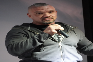 Ein Mann in einer schwarzen Kapuzenjacke, einem schwarzen Anzug und einem weißen Hemd mit zurückgekämmtem Haar hält ein Mikrofon und steht selbstbewusst vor einer Wand.