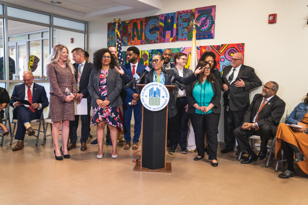 Eine Gruppe von Menschen steht vor einem Rednerpult mit einem Mikrofon und einer Tafel mit der Aufschrift "Sancho-Stadtrat tritt dem Stadtrat bei", im Hintergrund sind andere Menschen auf Stühlen und auf dem Boden sitzend und stehend, Flaggen, eine Tafel, ein Feuerlöscher und Deckenlampen an der Rückwand.