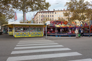 Eine belebte Stadtstraße mit einer Zebrastreifen und einem Karussell in der Mitte, mit Menschen, Buden, Bäumen, Gebäuden mit Fenstern, Bannern, Laternenpfählen und einer Ampel unter einem bewölkten Himmel.