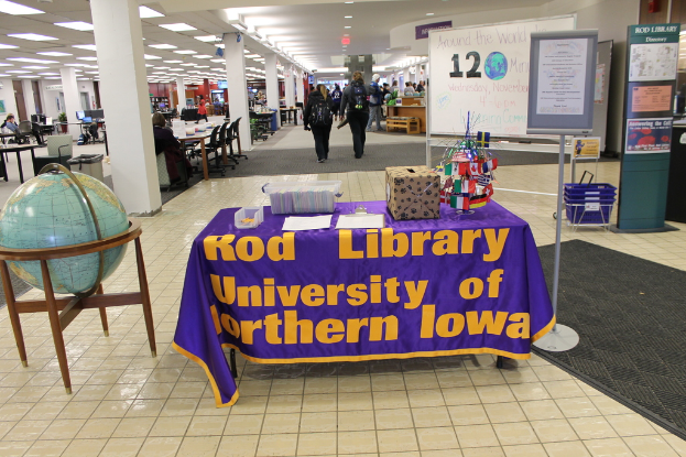 Eine Ausstellung mit einem Tisch, auf dem verschiedene Landesflaggen, eine Box, Papiere und ein Banner sowie ein Globus links und ein Anschlagbrett rechts mit einigen Menschen in der Nähe und mehreren Tischen und Stühlen links zu sehen sind.