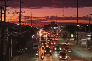 Eine Stadtstra├če bei Nacht voller Verkehr, darunter Autos und Motorräder, unter Straßenlaternen mit Gebäuden, Bäumen und Strommasten mit Drähten im Hintergrund und einem bewölkten Himmel.