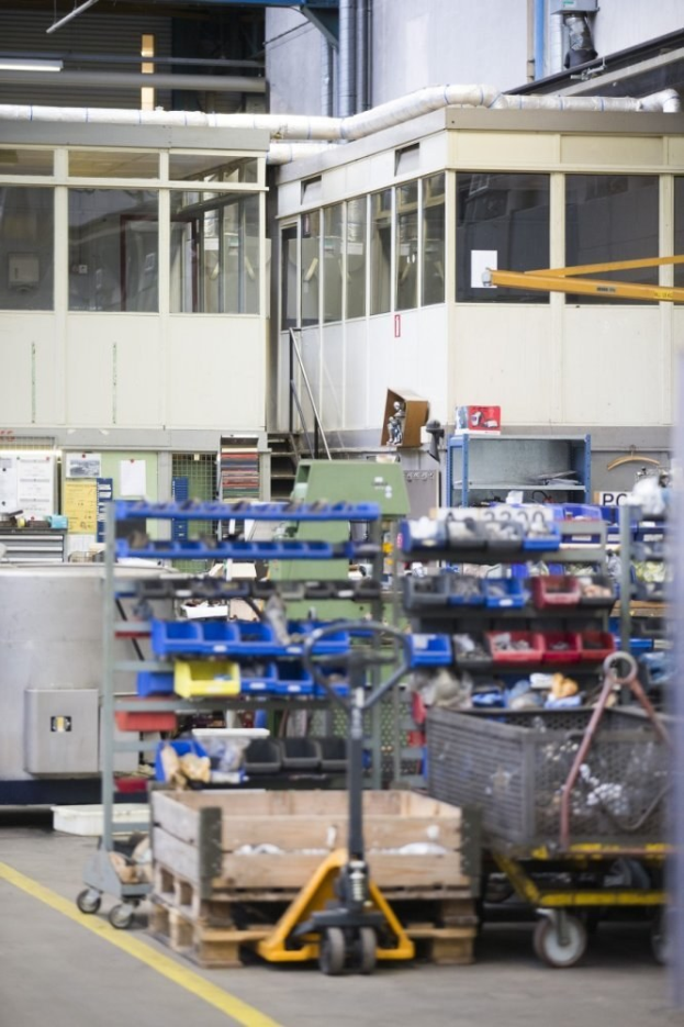 Ein großes Lagerhaus voller verschiedener Ausrüstungen wie Wagen, Schränken und einer Kiste, mit einer Wand aus Glasfenstern und Rohren im Hintergrund.