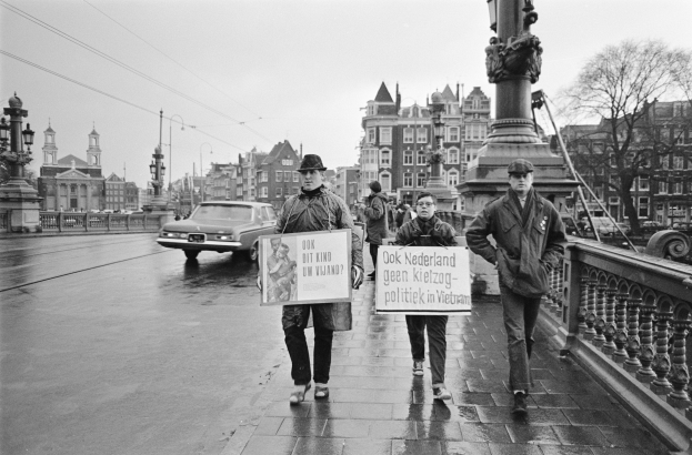 Eine Gruppe von Menschen überquert eine Brücke und hält Schilder in den Händen, während Fahrzeuge auf der Straße darunter fahren und Gebäude, Laternenpfähle, Bäume und ein klarer Himmel im Hintergrund zu sehen sind.