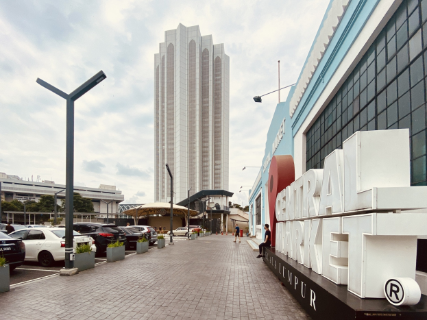 Ein belebter zentraler Markt in Kuala Lumpur, Malaysia, mit hohen Gebäuden, Straßenlaternen, Straßenlaternen, Kraftfahrzeugen, Fußgängern, Namensschildern, Bäumen und einer bewölkten Himmel.