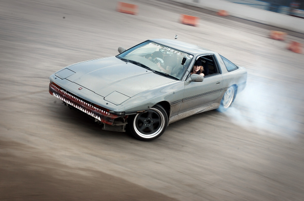 Eine Person in einem Helm fährt ein Auto auf einer von orangefarbenen Objekten umgebenen Strecke, mit einem verrauschten Hintergrund, der hohe Geschwindigkeit andeutet.
