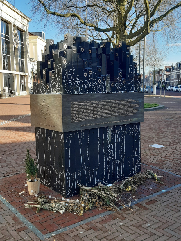Denkmal in einem Stadtplatz für Holocaust-Opfer, mit einem Baum davor, einer Topfpflanze, verstreuten trockenen Blättern und Gebäuden, Fahrzeugen, Masten und Bäumen im Hintergrund.