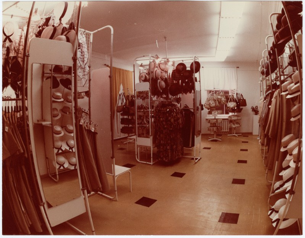 Altes Foto eines Kleiderladens mit Kleiderstangen, Hutständern, einem Spiegel, einem Stuhl, einem Tisch mit Gegenständen, Vorhängen und Deckenleuchten.