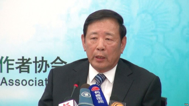 Ein Mann im Anzug und Krawatte sitzt an einem Tisch mit Mikrofonen und hält ein Plakat in den Händen.