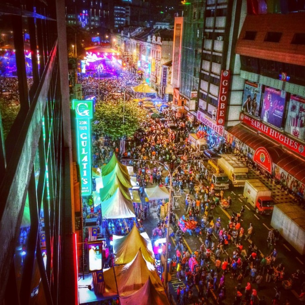 Eine belebte KarnevalsSzene mit einer großen Menschenmenge auf der Straße, umgeben von Zelten, Geschäften, Gebäuden und Bannern, mit zahlreichen Lichtern im Hintergrund.