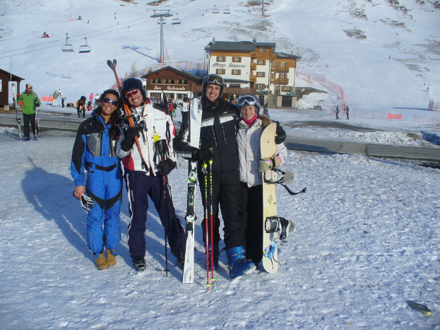 Vier Personen posieren für ein Foto mit Skating Pads und Werkzeugen, mit Gebäuden und anderen Menschen im Hintergrund, vor einem schneebedeckten Berg.