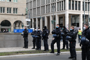 Eine Gruppe von Polizisten in schwarzen Uniformen und Masken steht vor einem Glasfenstergebäude mit Säulen in Berlin, Deutschland, einige halten Helme, mit Laternen, Verkehrszeichen, Schildern mit Text, einer Statue, anderen Menschen und grasbewachsenem Boden im Hintergrund.
