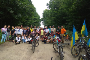 Eine Gruppe von Menschen mit Fahrrädern auf einem Schotterweg umgeben von Bäumen unter einem klaren blauen Himmel, einige tragen Helme und Taschen.