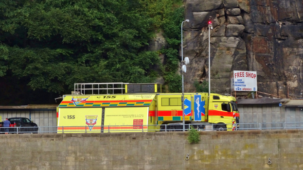 Ambulanz auf der Seite einer Straße neben einer Klippe geparkt, mit verschiedenen Gegenständen wie einer Wand, einem Geländer, Pfosten, Lichtern und Schildern mit Text im Vordergrund und Bäumen und einem Auto im Hintergrund.